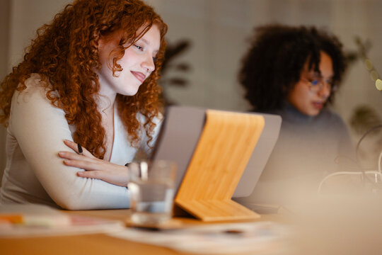 Two young women collaborate on a project using a tablet and laptop in a modern office setting.
