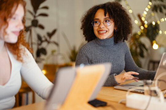Two young women collaborate on a project using laptops at a wooden table, with warm string lights in the background.