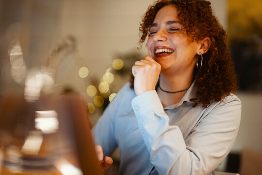 A young woman with curly hair laughs heartily while sitting at a table, her eyes closed in amusement.