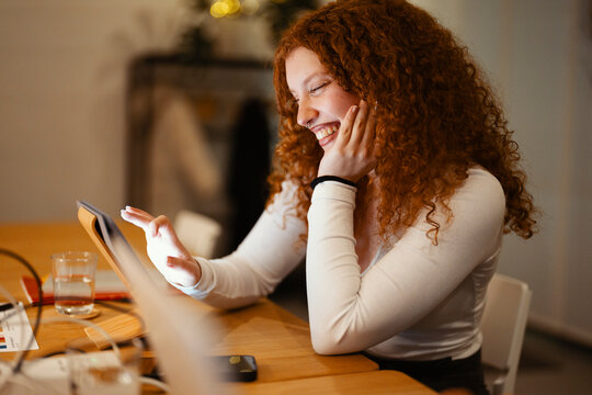 A young woman with curly red hair laughs while looking at a tablet computer. She is sitting at a wooden table with a glass of water and other items.