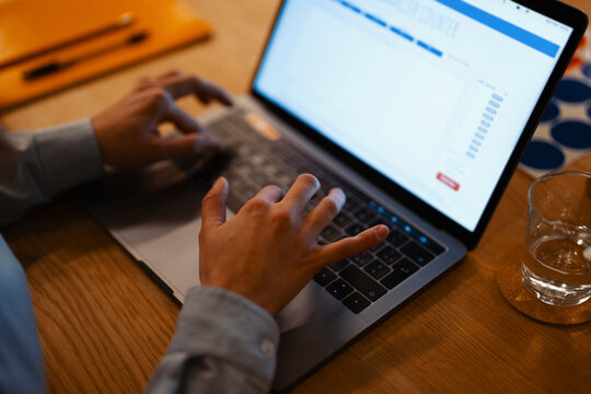 A person's hands are typing on a laptop keyboard, with a glass of water and a notebook on a wooden desk.