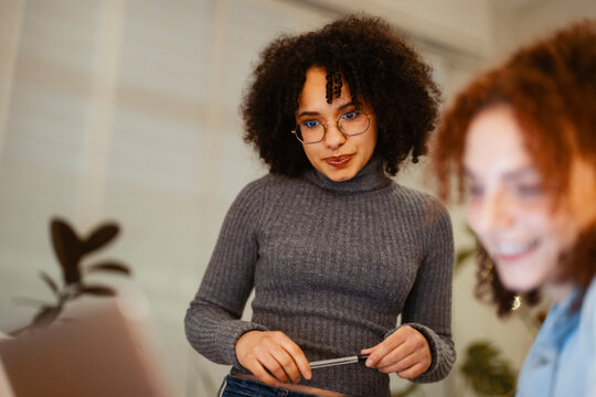 Two young women collaborate on a project, one focused on a laptop while the other holds a pen, suggesting teamwork and shared learning.