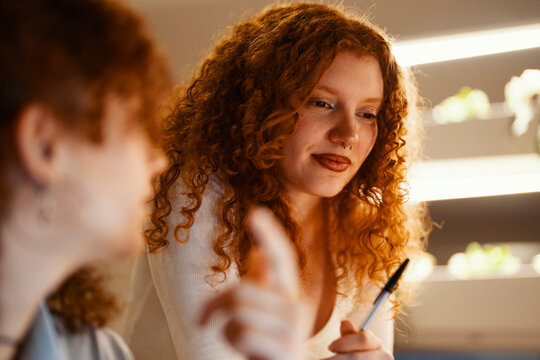 Two young women with curly red hair are engaged in a discussion, one gesturing with a pen while the other listens intently.