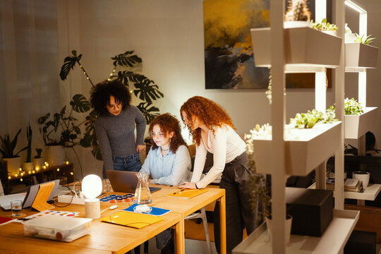Three young women collaborate around a laptop on a wooden table, surrounded by plants and warm lighting, suggesting a creative or study session.