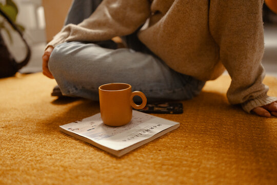 A person sits cross-legged on a yellow rug, holding a mug and a magazine. The scene evokes a sense of relaxation and quiet contemplation.