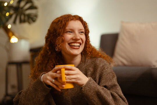 A young woman with curly red hair laughs while holding a yellow mug. She is wearing a cozy sweater and sitting in a dimly lit room with string lights.