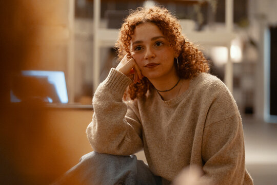 A young woman with curly red hair rests her head on her hand, looking thoughtfully into the distance while sitting indoors.
