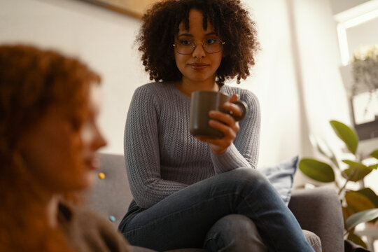 Two women are having a conversation while one holds a mug. They are sitting in a living room with soft lighting.