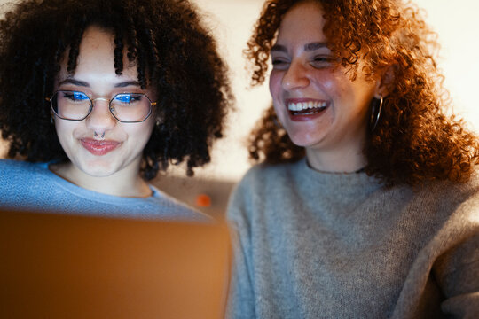 Two young women with curly hair share a laugh while looking at a laptop screen, enjoying a moment of connection and shared amusement.