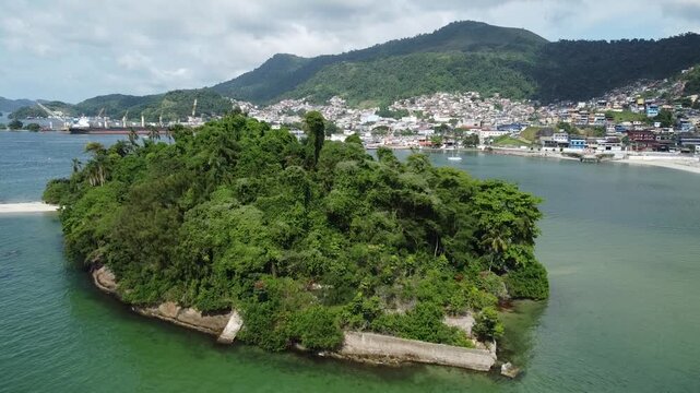 Drone rises behind the small tropical Island, covered in dense Atlantic rainforest, Ilha do Coqueiro in the Bay of Angra dos Reis, and reveal the Angra dos Reis centre behind it