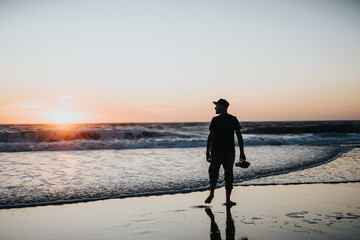 A man walks barefoot along the wet sand at sunset, holding his shoes and gazing toward the horizon as gentle waves roll in. A tranquil, contemplative scene of travel and seaside relaxation.