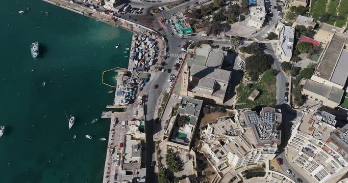 Aerial view of Marsaskala Parish Church, boats in harbor, and buildings displaying a blend of textures and tones, Marsaskala, Wied il-Ghajn, Malta.