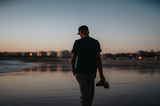 A man walks along a quiet, wet shoreline at dusk, holding a pair of sneakers. The horizon glows softly, and distant buildings reflect in the water, creating a serene, contemplative scene.