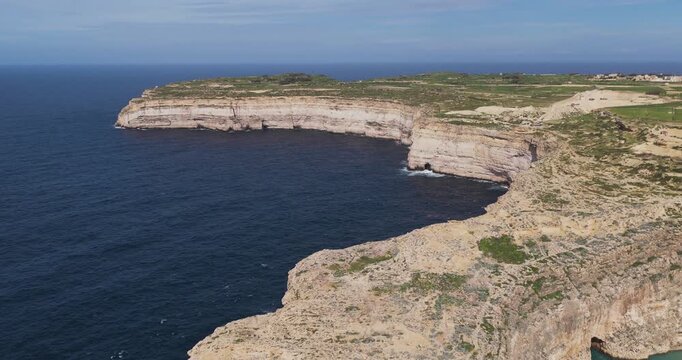 Aerial view of the rugged coastline featuring beige rocky cliffs meeting the dark blue sea in Dwejra Bay, Gozo Island, Malta.