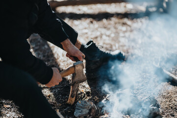 Obraz premium A person chops wood with an axe near a crackling campfire outdoors. A simple scene of hands, wood, and smoke in a natural setting.