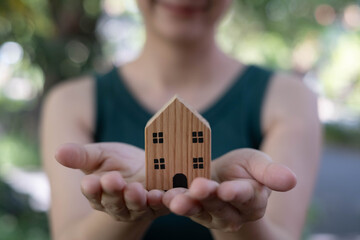 Woman holding a small wooden house in her hands outdoors, symbolizing home care, property...