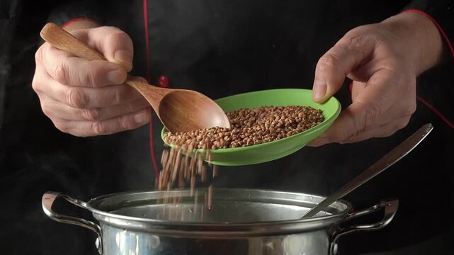 Chef pours buckwheat from a green bowl into a stainless steel pot using a wooden spoon, steam rising in a kitchen setting with dark background