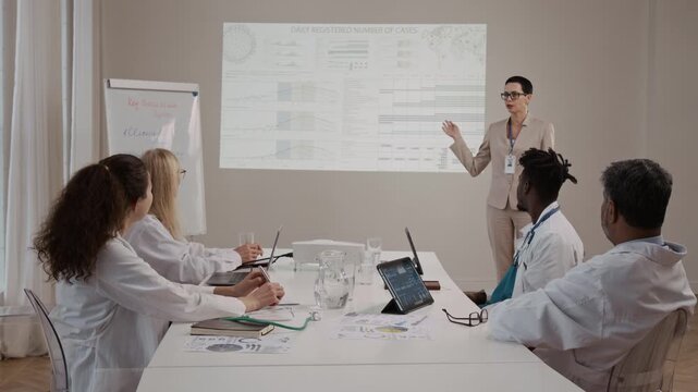 Diverse team of medical specialists in lab gowns sitting at large white table listening to Caucasian mature speaker in pantsuit giving presentation on daily registered number of cancer cases