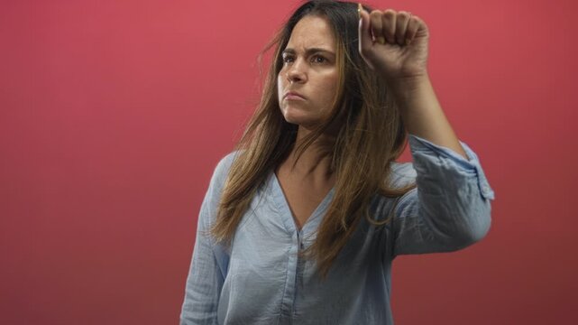 Woman raises fist and holds up her hand in a red studio with focused gaze and slight forward motion toward camera; defiance anger.