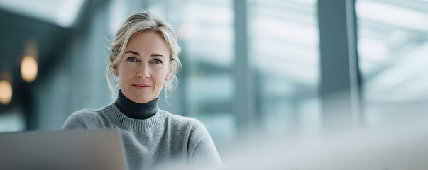 Mature blonde female executive smiles confidently in a bright modern office setting, laptop visible, concept for corporate leadership, professional development and business success