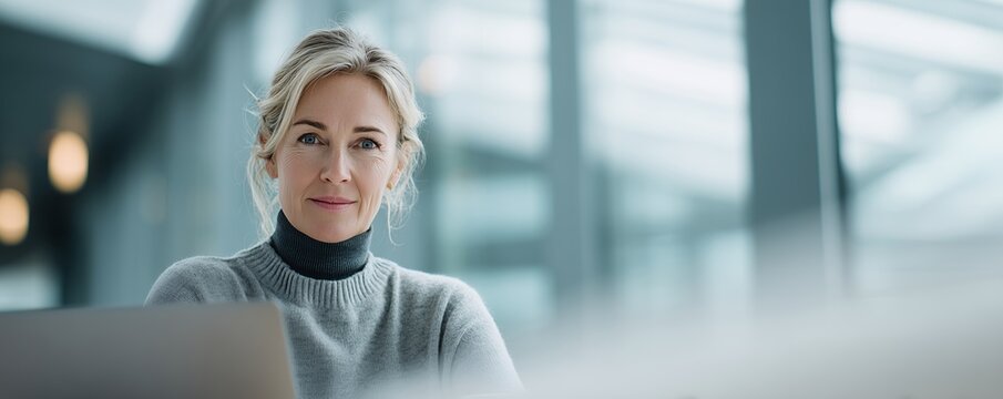 Mature blonde female executive smiles confidently in a bright modern office setting, laptop visible, concept for corporate leadership, professional development and business success