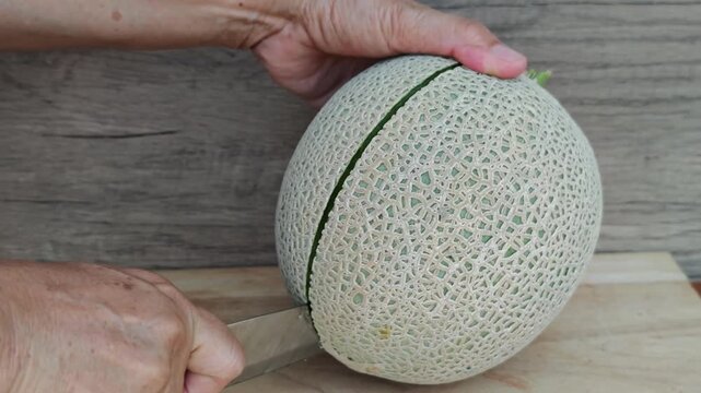 Close-up of a hand slicing a cantaloupe melon in half on a wooden cutting board.