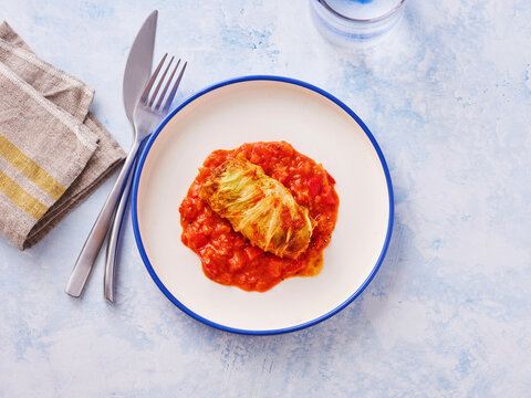 A stuffed cabbage roll is served on a white plate with tomato sauce. Cutlery is placed beside the plate on a table. The setting is bright and inviting, suitable for a meal