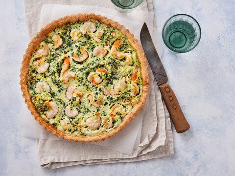A shrimp and spinach quiche is placed on a light surface. Next to it is a knife. There are two glasses beside the quiche. The setting shows a clean kitchen space
