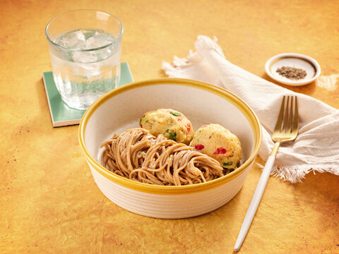 A nourishing meal featuring soba noodles and two vegetable dumplings served in a bowl on a golden-yellow table. A glass of ice water, a golden fork, and a small dish of spices complete the setting