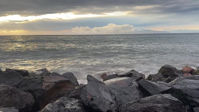 Ocean waves crash onto black volcanic rocks, creating white spray and foam beneath dramatic storm clouds at sunset. Natural coastal seascape with shimmering water and wide horizon