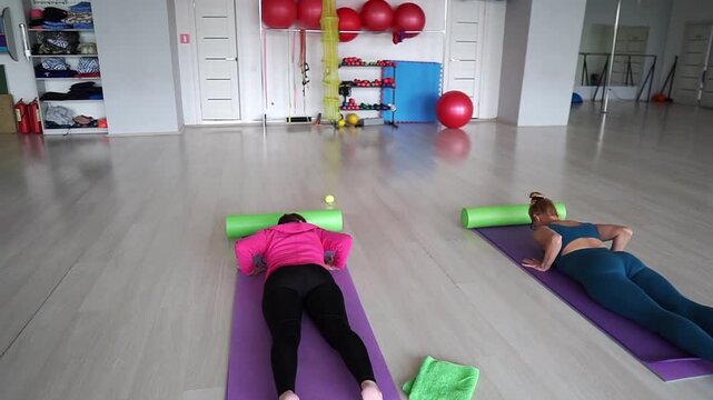 Two women perform prone back extension exercise on mats in calm studio setting, with focused breathing and gentle rehabilitation under guidance of trainer. Fitness, stretching, rehabilitation concepts