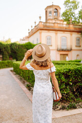 Fototapeta premium A woman stands outdoors in a garden with bushes. She wears a hat and smiles as she adjusts it. A building is visible behind her during late afternoon light.