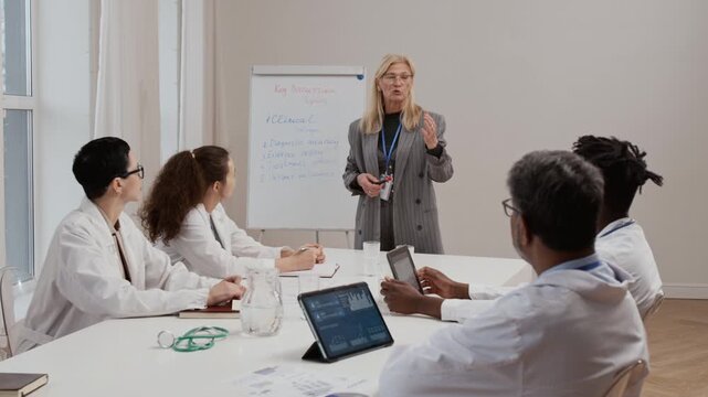 Caucasian blonde mature health professional giving speech on treatment options to multiethnic group of physicians sitting at large table in conference room