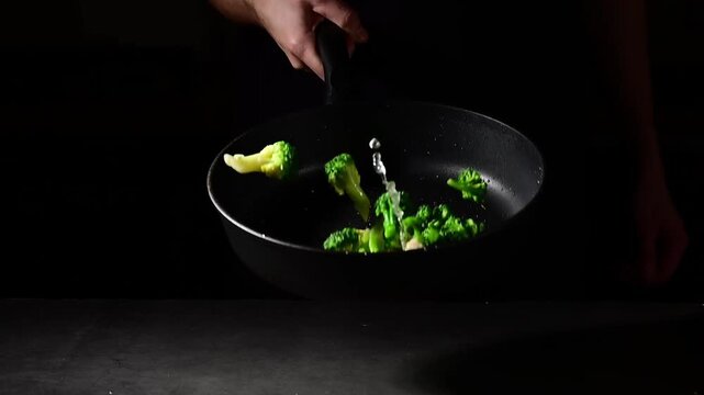 Chef tossing broccoli florets in hot cast iron skillet with wrist flick technique, slow motion capture of vegetable arc and oil droplets in professional kitchen environment