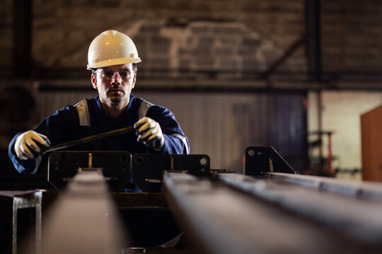 Industrial worker measuring a metal component in a factory workshop while wearing safety helmet and gloves, ensuring precision, quality control, and workplace safety in manufacturing process.