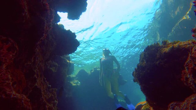 still view of slow motion of a skinny slim Caucasian female blond free diver wearing mask and snorkel swimming accenting up facing to the camera to the surface around coral reef cliffs in the blue oce