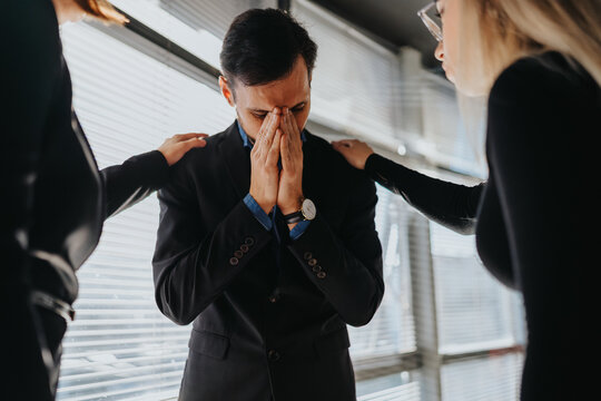 A man in a suit covers his face with folded hands while two colleagues place hands on his shoulders. The office scene conveys support and empathy during a stressful moment.