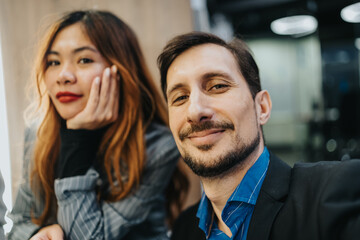 A man smiles at the camera while a woman rests her chin on her hand in the background of a contemporary office.