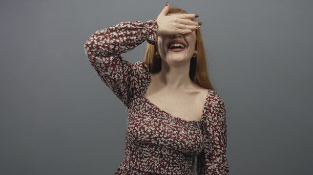 Woman covering her eyes with hand while smiling in a studio, young redhead in a floral dress showing shoulder and hoop earrings; playful joy.