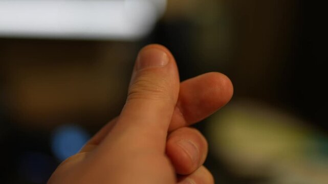 Close-up of a hand making a "finger heart" gesture. Soft, warm lighting illuminates the skin. The background is heavily blurred with dark tones, an out-of-focus white light source