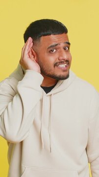 I cant hear you. What. Indian man trying hear you, looking confused and frowning, keeping arm near ear for louder voice, asking to repeat, to hear information, deafness. Young guy on yellow background