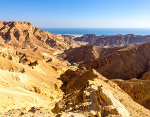 Expansive desert landscape with tan mountains and a glimpse of the ocean. Bright sunlight and blue sky. View from a rocky ridge