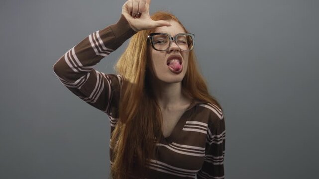 Redhead woman wearing brown striped sweater sticks out tongue and makes l with hand to forehead in studio; cheeky defiance.