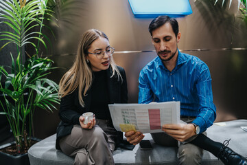 Two colleagues review a document on a sofa in a modern lobby. They discuss the report in a bright, plant-filled space.