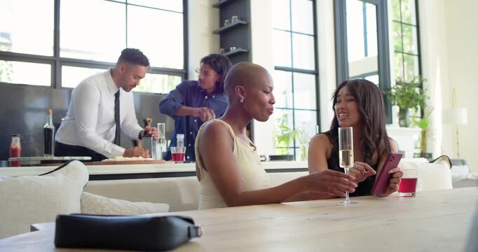 Diverse female friends leaning at table in apt sharing phone clinking flute men prepping drinks