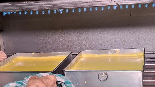 An elderly woman's hand places a pan of Lapis Legit batter into the oven, next to another pan already inside.