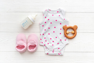 Baby bodysuits and eco friendly accessories - booties, wooden rattle toy and baby bottle with milk on white wooden background, top view