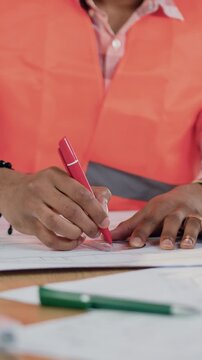 Unrecognizable purposeful skilled man-constructor in protective vest sitting at workplace in engineering workroom and working with blueprint,he uses pen and protractor to draw line