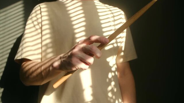 Young man hands skillfully spinning, drop drumstick, practicing his rhythm and dexterity. Dramatic shadows from window blinds create striking visual pattern on t shirt. Drummer spinning drumstick