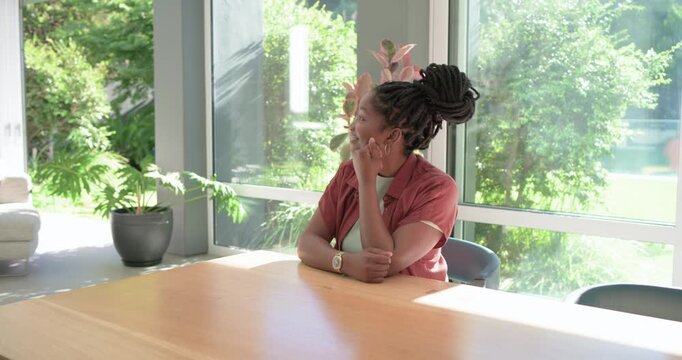 Adult African woman sitting at table by glass windows, gazing out, turning to camera, greeting